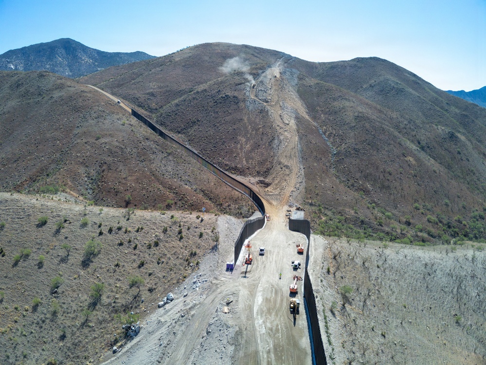 An aerial photograph shows active construction of a large border wall/barrier traversing rugged, arid mountainous terrain. The wall extends from the foreground valley up and over steep hillsides into the distance. Multiple pieces of heavy construction equipment and vehicles are visible along a cleared dirt access road running parallel to the barrier.