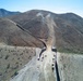 An aerial photograph shows active construction of a large border wall/barrier traversing rugged, arid mountainous terrain. The wall extends from the foreground valley up and over steep hillsides into the distance. Multiple pieces of heavy construction equipment and vehicles are visible along a cleared dirt access road running parallel to the barrier.