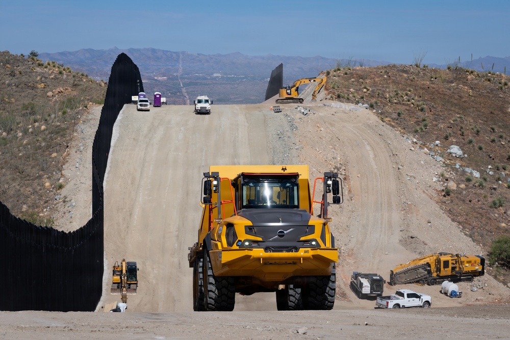 A large-scale construction project is underway along a tall border barrier made of dark steel bollards. Heavy construction equipment, including a prominent yellow articulated dump truck in the foreground, is actively working on a graded dirt road running parallel to the wall. The terrain is arid and hilly, with mountains visible in the background under a clear blue sky.