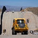 A large-scale construction project is underway along a tall border barrier made of dark steel bollards. Heavy construction equipment, including a prominent yellow articulated dump truck in the foreground, is actively working on a graded dirt road running parallel to the wall. The terrain is arid and hilly, with mountains visible in the background under a clear blue sky.