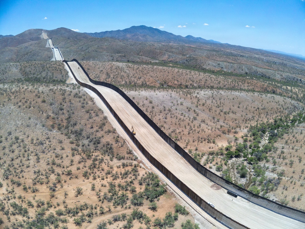 An aerial photograph showing a long border barrier or security fence stretching across an arid, desert landscape with sparse vegetation. The structure runs diagonally from foreground to background, flanked by a cleared dirt patrol road on one or both sides. Mountains are visible in the background under a clear blue sky.
