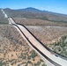 An aerial photograph showing a long border barrier or security fence stretching across an arid, desert landscape with sparse vegetation. The structure runs diagonally from foreground to background, flanked by a cleared dirt patrol road on one or both sides. Mountains are visible in the background under a clear blue sky.