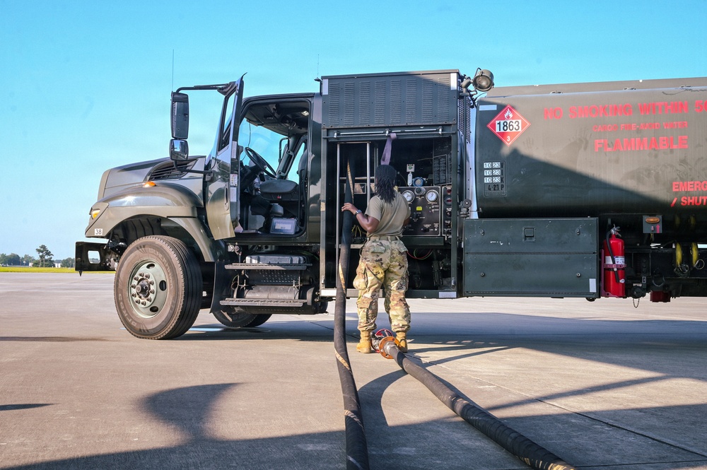A military service member in camouflage uniform is operating a large fuel tanker truck on what appears to be an airfield tarmac. The vehicle is a military-green fuel bowser with hazmat placards and flammable material warnings. The service member is handling a large fuel hose connected to the tanker's pump control panel.