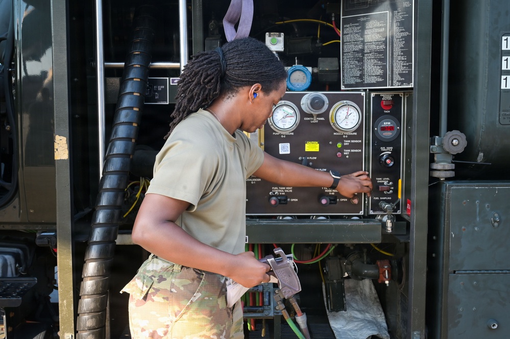 A female military service member in U.S. Army combat uniform is operating a fuel distribution or pumping control panel on a military vehicle. She is wearing a tan t-shirt and multicam/OCP pattern pants, with blue ear protection inserted, and is manipulating controls on the panel with one hand while holding equipment in the other. The control panel displays gauges, indicators, and labeled valves consistent with a military fuel handling system.