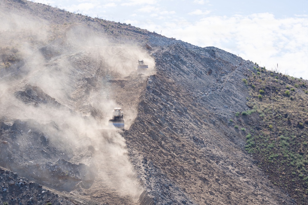 The image shows an active open-pit mining or large-scale earthmoving operation on a steep hillside. Two heavy construction machines, likely bulldozers or wheel loaders, are visible working on a cut slope, generating significant dust clouds. The terrain shows exposed rock and soil with sparse desert vegetation on the undisturbed portions of the hill.