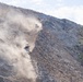 The image shows an active open-pit mining or large-scale earthmoving operation on a steep hillside. Two heavy construction machines, likely bulldozers or wheel loaders, are visible working on a cut slope, generating significant dust clouds. The terrain shows exposed rock and soil with sparse desert vegetation on the undisturbed portions of the hill.