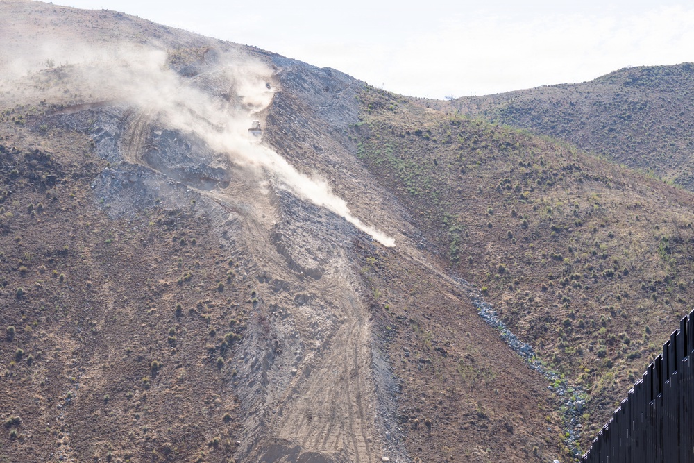 The image shows heavy construction or grading activity on a steep hillside, with visible dust clouds rising from machinery operating on a cleared dirt path cut into the slope. A dark vertical barrier structure is visible in the lower right corner of the frame. The terrain is arid and semi-vegetated with sparse shrubs and small trees.