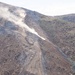 The image shows heavy construction or grading activity on a steep hillside, with visible dust clouds rising from machinery operating on a cleared dirt path cut into the slope. A dark vertical barrier structure is visible in the lower right corner of the frame. The terrain is arid and semi-vegetated with sparse shrubs and small trees.