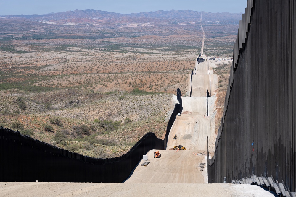 The image shows a large border wall under active construction, extending into the distance across an arid desert landscape. The wall consists of tall, dark vertical steel bollards and appears to be newly installed, with construction equipment and a dirt access road visible alongside it. The terrain is characteristic of a southwestern desert region with sparse vegetation and mountain ranges in the background.