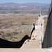 The image shows a large border wall under active construction, extending into the distance across an arid desert landscape. The wall consists of tall, dark vertical steel bollards and appears to be newly installed, with construction equipment and a dirt access road visible alongside it. The terrain is characteristic of a southwestern desert region with sparse vegetation and mountain ranges in the background.