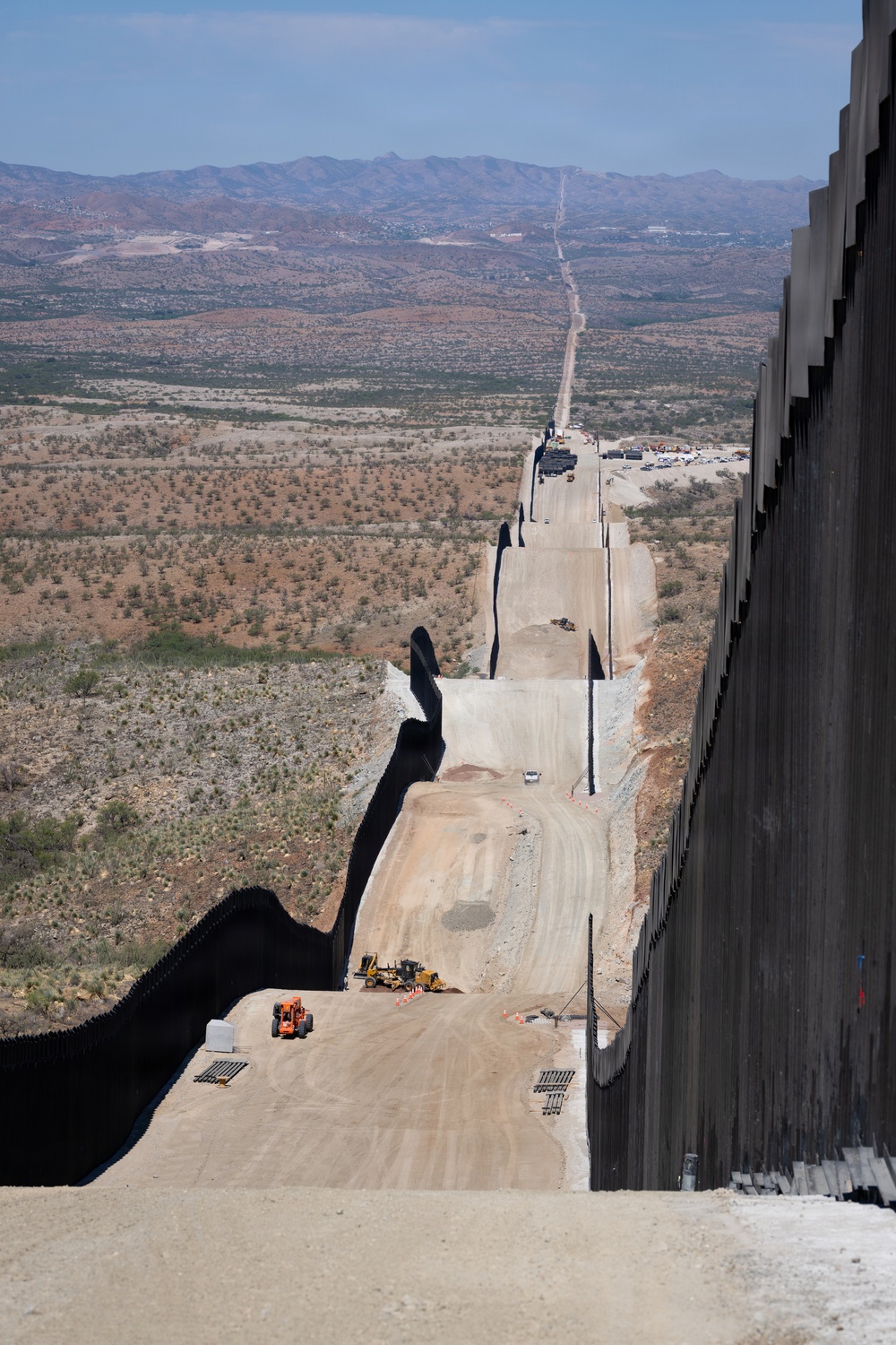 An aerial or elevated view of a large border wall under active construction, stretching into the distance across arid desert terrain. The wall consists of tall dark steel bollard-style panels running parallel, with a graded patrol road between them. Construction equipment and vehicles are visible along the corridor, with mountainous landscape visible in the background.