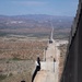An aerial or elevated view of a large border wall under active construction, stretching into the distance across arid desert terrain. The wall consists of tall dark steel bollard-style panels running parallel, with a graded patrol road between them. Construction equipment and vehicles are visible along the corridor, with mountainous landscape visible in the background.