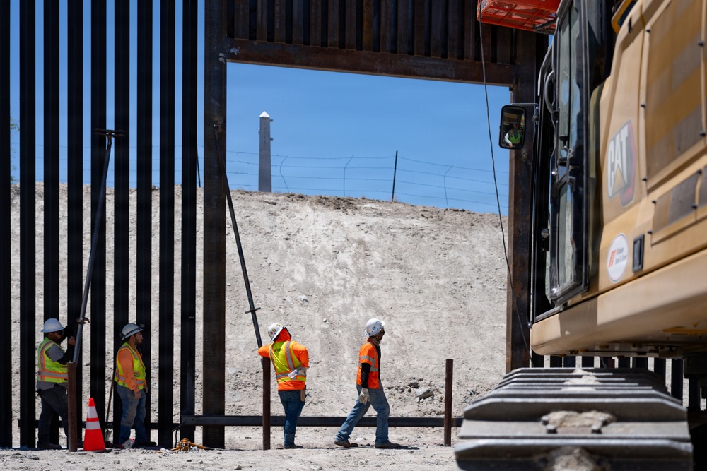 Construction workers are actively building or reinforcing a large steel border barrier/wall structure. Four workers in high-visibility orange safety vests and white hard hats are working alongside a CAT excavator on a cleared, sandy construction site. A secondary fence with barbed wire is visible in the background through an opening in the primary wall.