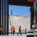 Construction workers are actively building or reinforcing a large steel border barrier/wall structure. Four workers in high-visibility orange safety vests and white hard hats are working alongside a CAT excavator on a cleared, sandy construction site. A secondary fence with barbed wire is visible in the background through an opening in the primary wall.