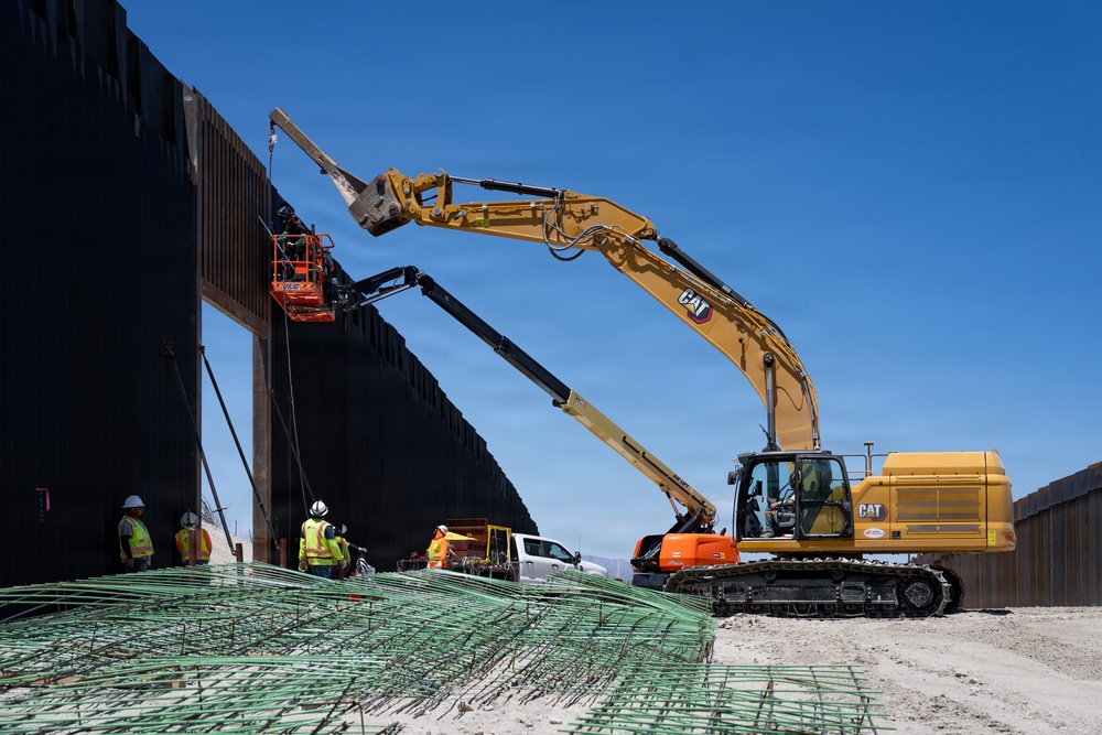 A large-scale construction site featuring a tall dark steel barrier wall being erected using heavy machinery. A yellow CAT excavator with an extended boom arm is actively working alongside a red aerial lift platform with workers. Green rebar bundles are staged in the foreground, and multiple workers in high-visibility safety gear are present on site.