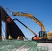 A large-scale construction site featuring a tall dark steel barrier wall being erected using heavy machinery. A yellow CAT excavator with an extended boom arm is actively working alongside a red aerial lift platform with workers. Green rebar bundles are staged in the foreground, and multiple workers in high-visibility safety gear are present on site.