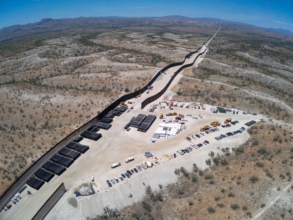 An aerial drone photograph showing an active border wall construction site in an arid, desert landscape. A dark vertical barrier wall extends from the foreground into the distance along a ridgeline, with a large staging area containing construction materials, heavy equipment, and vehicles on one side. Mountains are visible in the background under clear blue skies.