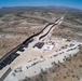 An aerial drone photograph showing an active border wall construction site in an arid, desert landscape. A dark vertical barrier wall extends from the foreground into the distance along a ridgeline, with a large staging area containing construction materials, heavy equipment, and vehicles on one side. Mountains are visible in the background under clear blue skies.