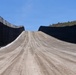 The image shows a dirt patrol road running between two parallel sections of tall steel border barrier panels. The barriers are dark-colored vertical steel bollards extending into the distance over hilly terrain. The scene appears to be a recently constructed or maintained border infrastructure corridor in an arid, semi-vegetated landscape.
