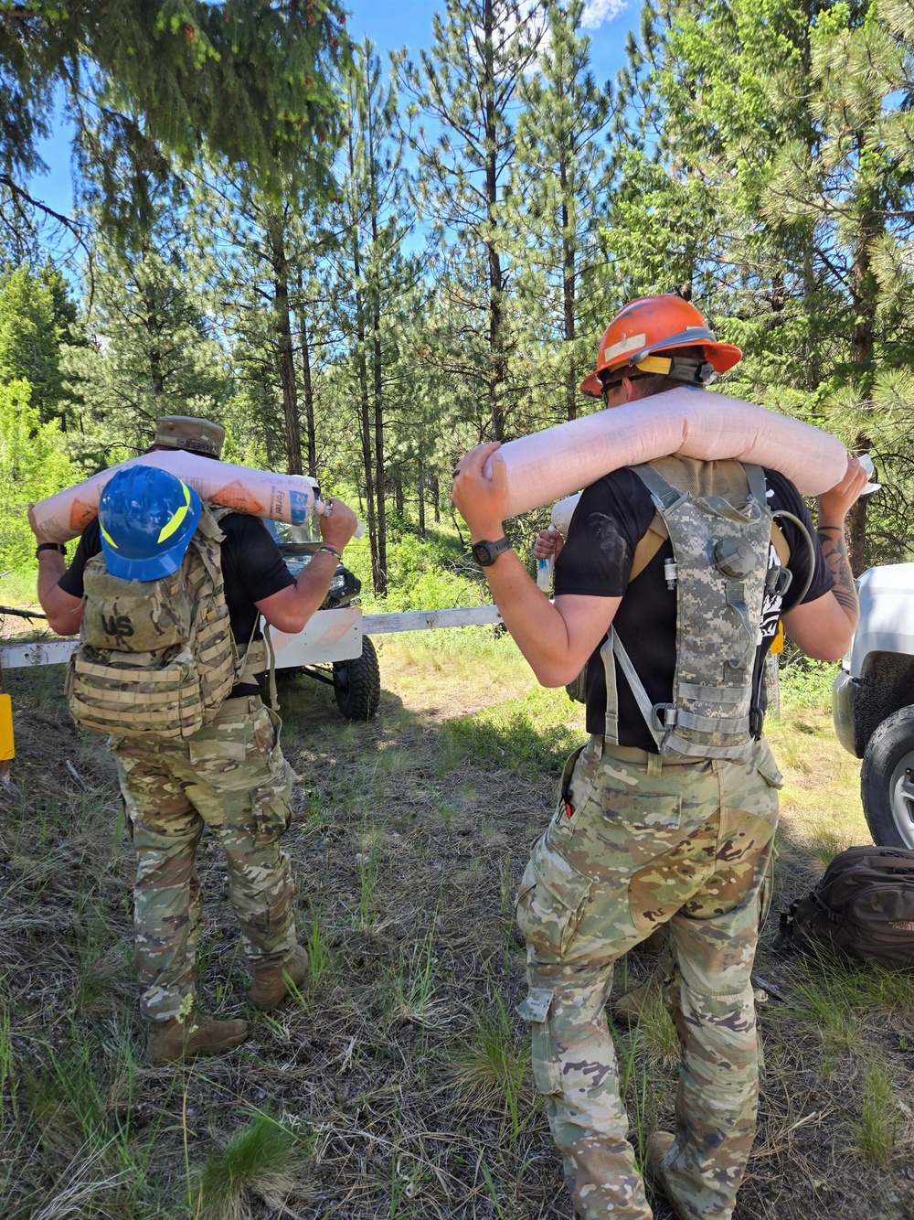 Two individuals in US Army camouflage (OCP pattern) uniforms are carrying large inflatable or padded cylindrical objects on their shoulders in a forested outdoor setting. Both wear tactical gear including plate carriers/vests and hard hats, suggesting a wildfire support or civil assistance mission. Vehicles and a trailer are visible in the background among pine trees.