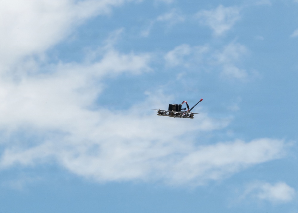 A small racing or FPV (First Person View) drone is captured mid-flight against a partly cloudy blue sky. The drone appears to be a custom-built quadcopter with visible components including what appears to be a battery and antenna. The image was taken from below, looking upward, with the drone positioned in the center-right of the frame.