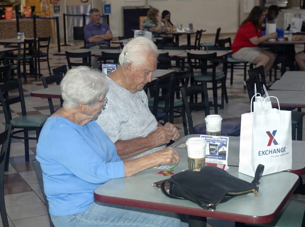 Two elderly individuals, a man and a woman, are seated at a table in what appears to be a military base Exchange food court or cafeteria. They appear to be playing a game, likely bingo, with colored markers on a card. A white 'Exchange' shopping bag, coffee cups, and a black purse are visible on the table.