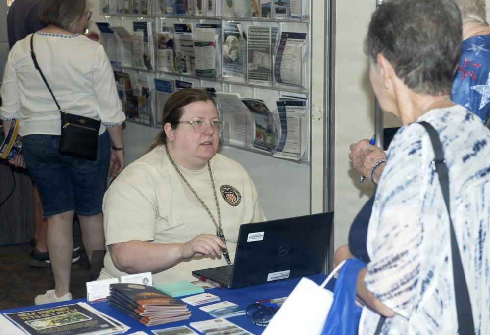 A woman seated at an information or outreach booth is speaking with a visitor at what appears to be a public event or fair. The booth worker wears a cream-colored shirt with a medallion/badge necklace and has a Dell laptop open in front of her. The table is covered with brochures, booklets, and informational materials, and a rack of pamphlets is visible in the background.