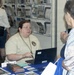 A woman seated at an information or outreach booth is speaking with a visitor at what appears to be a public event or fair. The booth worker wears a cream-colored shirt with a medallion/badge necklace and has a Dell laptop open in front of her. The table is covered with brochures, booklets, and informational materials, and a rack of pamphlets is visible in the background.