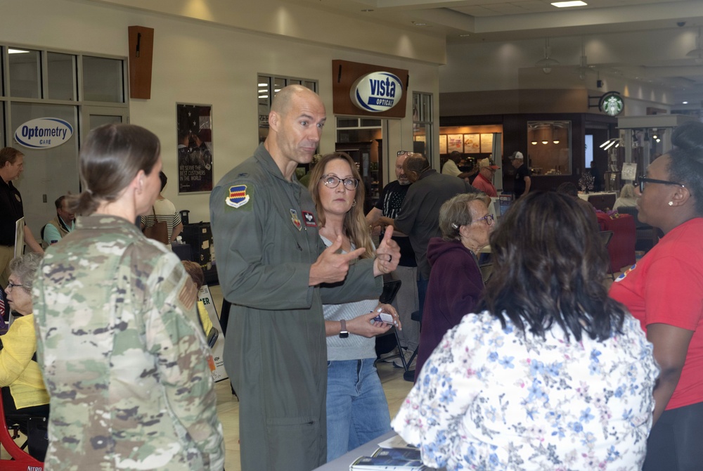 A military community event or health fair is taking place inside what appears to be a military base exchange or commissary facility. A U.S. Air Force officer in a flight suit and a soldier in Army combat uniform are interacting with civilians at what appears to be an informational or outreach event. The setting includes retail stores such as Vista Optical, an Optometry Care clinic, and a Starbucks in the background.