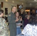 A military community event or health fair is taking place inside what appears to be a military base exchange or commissary facility. A U.S. Air Force officer in a flight suit and a soldier in Army combat uniform are interacting with civilians at what appears to be an informational or outreach event. The setting includes retail stores such as Vista Optical, an Optometry Care clinic, and a Starbucks in the background.