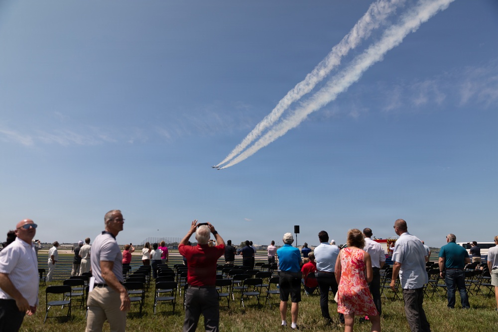 An outdoor airshow event where spectators are gathered on a grassy field watching aircraft perform aerial maneuvers. Two jets are visible in the sky leaving parallel white smoke trails as they fly in close formation. Folding chairs are arranged in rows suggesting an organized viewing area for the audience.