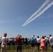 An outdoor airshow event where spectators are gathered on a grassy field watching aircraft perform aerial maneuvers. Two jets are visible in the sky leaving parallel white smoke trails as they fly in close formation. Folding chairs are arranged in rows suggesting an organized viewing area for the audience.