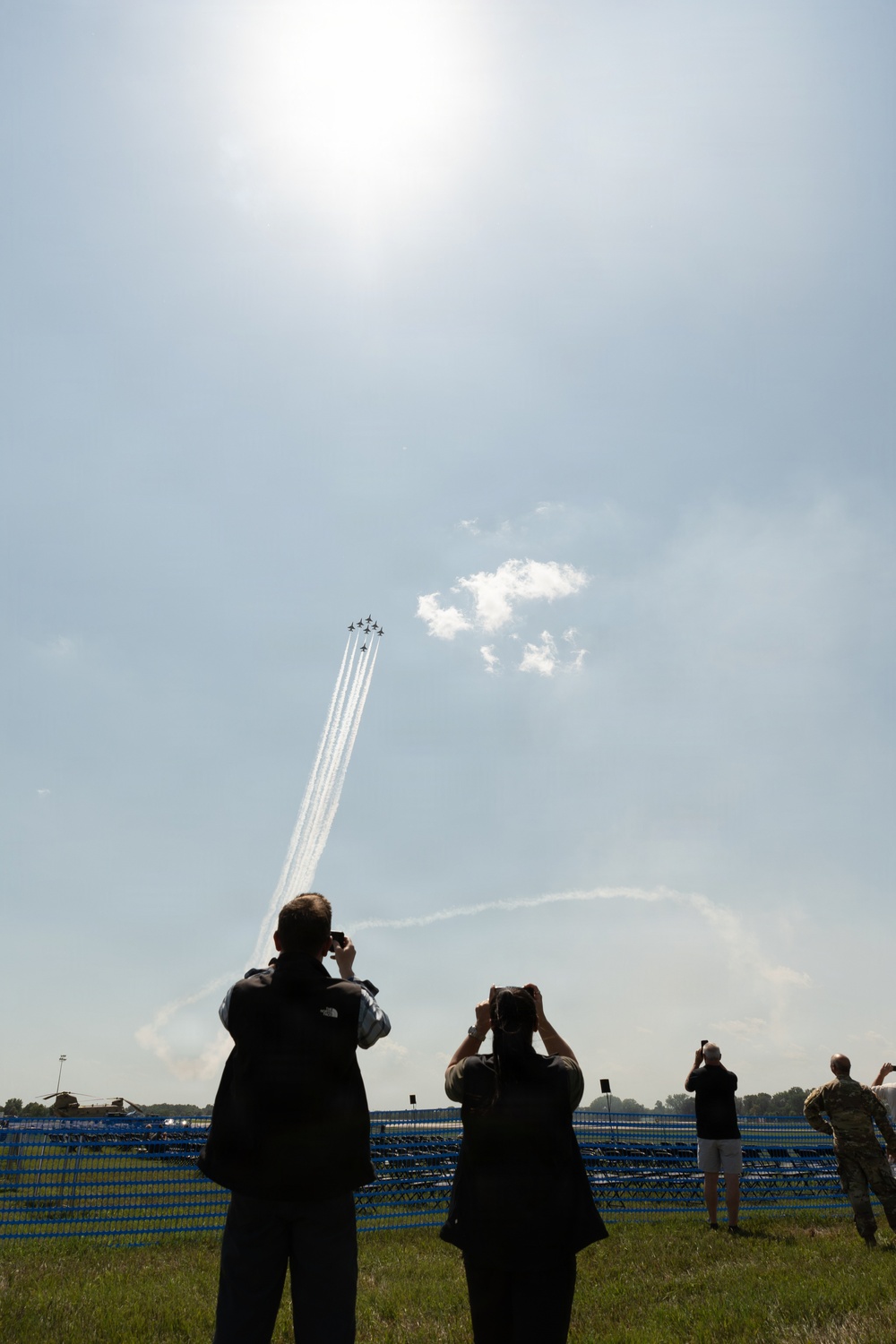 Spectators at an airshow watch and photograph a formation of jet aircraft performing an aerobatic maneuver, leaving white smoke trails as they climb steeply into a hazy sky. Multiple people are silhouetted against the bright sky, holding up cameras and smartphones to capture the display. In the background, bleacher seating, a fence perimeter, and what appear to be military helicopters are visible on the ground.