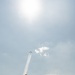 Spectators at an airshow watch and photograph a formation of jet aircraft performing an aerobatic maneuver, leaving white smoke trails as they climb steeply into a hazy sky. Multiple people are silhouetted against the bright sky, holding up cameras and smartphones to capture the display. In the background, bleacher seating, a fence perimeter, and what appear to be military helicopters are visible on the ground.