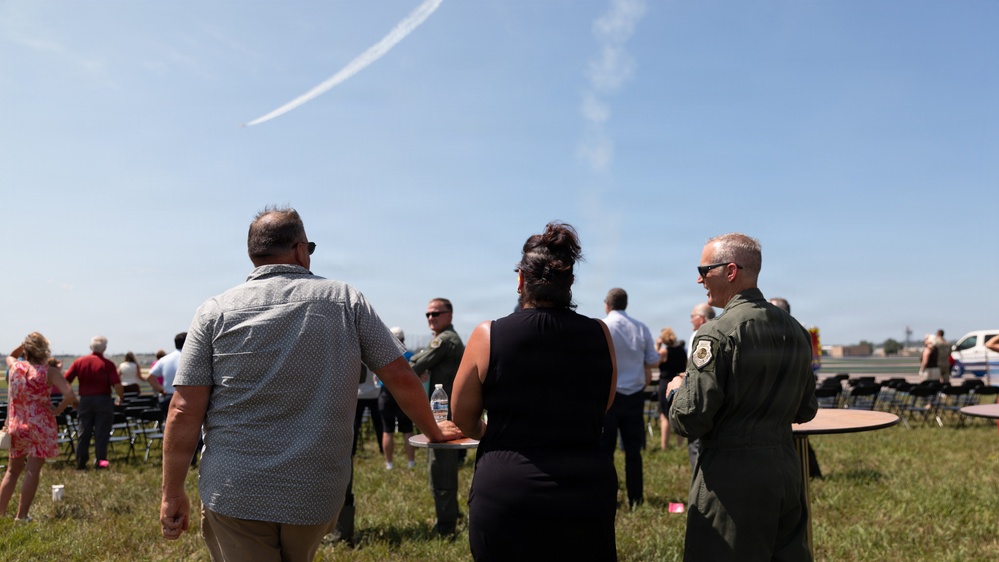 A group of people, including a military personnel in a flight suit, are gathered outdoors on a grassy area watching an aerial display. A white contrail arc is visible in the blue sky, likely from a jet performing a maneuver. The setting appears to be a military air show or ceremony with folding chairs arranged in the background.