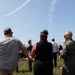 A group of people, including a military personnel in a flight suit, are gathered outdoors on a grassy area watching an aerial display. A white contrail arc is visible in the blue sky, likely from a jet performing a maneuver. The setting appears to be a military air show or ceremony with folding chairs arranged in the background.