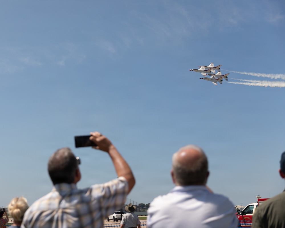 A crowd of spectators watches a precision aerial demonstration at an airshow, with multiple jet aircraft flying in tight formation. The jets, identifiable as U.S. Air Force Thunderbirds by their distinctive white and blue livery, are leaving white smoke trails as they fly in a diamond formation. Spectators in the foreground are photographing the display with smartphones.