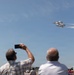 A crowd of spectators watches a precision aerial demonstration at an airshow, with multiple jet aircraft flying in tight formation. The jets, identifiable as U.S. Air Force Thunderbirds by their distinctive white and blue livery, are leaving white smoke trails as they fly in a diamond formation. Spectators in the foreground are photographing the display with smartphones.