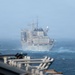 A large naval auxiliary or replenishment vessel is photographed at sea from the deck of another ship, likely during rough weather conditions. The distant ship appears to be a US Navy fleet replenishment oiler or combat stores ship, identifiable by its gray hull, superstructure, and characteristic replenishment-at-sea equipment. The image is taken from the foreground vessel's deck, with haze and sea spray reducing visibility.