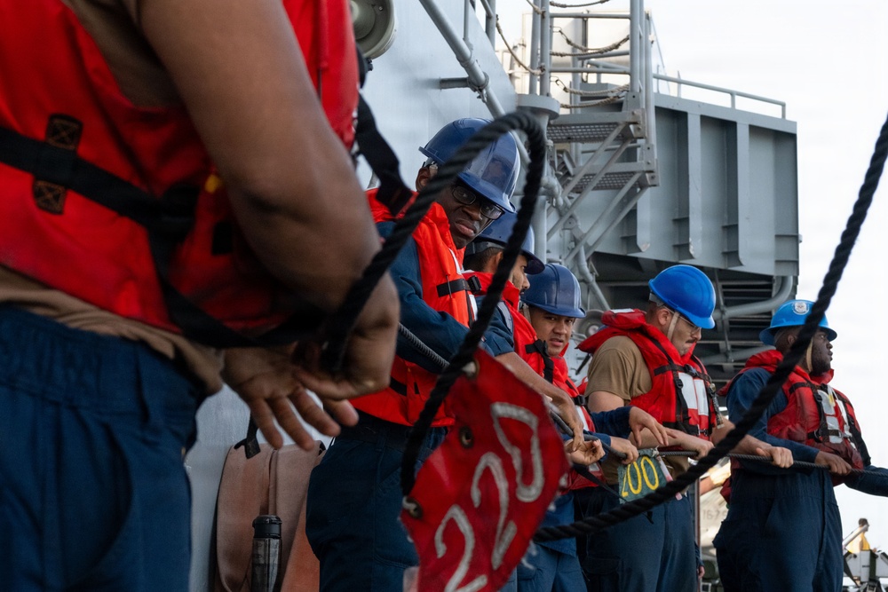 A group of naval personnel are working together on the deck of a military vessel, handling large mooring lines. The crew members are wearing standard safety equipment including blue hard hats and orange life vests over their uniforms. This appears to be a line-handling evolution, likely during a mooring or underway replenishment operation.