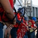 A group of naval personnel are working together on the deck of a military vessel, handling large mooring lines. The crew members are wearing standard safety equipment including blue hard hats and orange life vests over their uniforms. This appears to be a line-handling evolution, likely during a mooring or underway replenishment operation.
