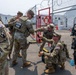 A group of U.S. National Guard soldiers in OCP (Operational Camouflage Pattern) uniforms are gathered near a secured perimeter gate topped with concertina wire. One soldier is crouching and appears to be operating or demonstrating a handheld electronic device to a standing colleague. The setting appears to be a security checkpoint or controlled access area with orange traffic cones and temporary structures visible in the background.