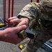 Two U.S. Army soldiers in MultiCam (OCP) camouflage uniforms are handling a SIG Sauer pistol magazine in an outdoor setting. One soldier appears to be passing or receiving the tan/coyote-colored magazine to/from the other. A U.S. flag patch and what appears to be a two-star general rank patch are visible on one soldier's sleeve.