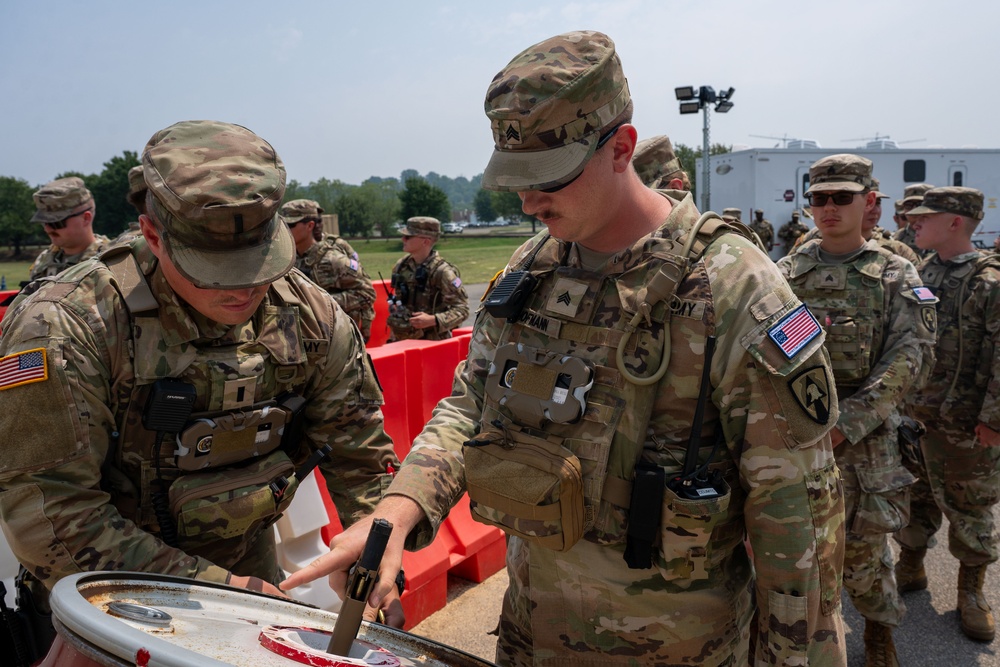 Two U.S. Army soldiers in OCP (Operational Camouflage Pattern) uniforms are examining an object on a surface, with one soldier pointing at it. They are wearing tactical vests and patrol caps, with rank insignia visible indicating Sergeant rank. A group of additional soldiers stands in the background near orange barriers and a white trailer.
