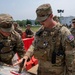 Two U.S. Army soldiers in OCP (Operational Camouflage Pattern) uniforms are examining an object on a surface, with one soldier pointing at it. They are wearing tactical vests and patrol caps, with rank insignia visible indicating Sergeant rank. A group of additional soldiers stands in the background near orange barriers and a white trailer.