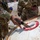 JTF-DC guardsmen prepare for patrol Two military personnel in MultiCam/OCP camouflage uniforms are inspecting or sampling the contents of a large metal drum or barrel with a red-ringed opening. One soldier is inserting what appears to be a handheld detection or sampling device into the drum's opening. The scene suggests a hazardous materials inspection, chemical detection, or field sampling exercise.