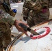 Two military personnel in MultiCam/OCP camouflage uniforms are inspecting or sampling the contents of a large metal drum or barrel with a red-ringed opening. One soldier is inserting what appears to be a handheld detection or sampling device into the drum's opening. The scene suggests a hazardous materials inspection, chemical detection, or field sampling exercise.
