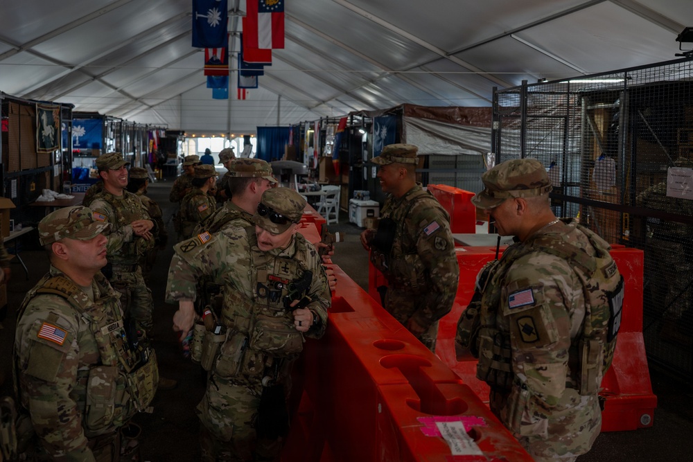 A group of U.S. Army soldiers in OCP (Operational Camouflage Pattern) uniforms are gathered inside a large tent or temporary structure. They appear to be at a staging or coordination area, congregating around orange plastic water-filled barriers. State flags are visible hanging from the tent ceiling, suggesting a multi-state National Guard or joint military operation.