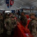 A group of U.S. Army soldiers in OCP (Operational Camouflage Pattern) uniforms are gathered inside a large tent or temporary structure. They appear to be at a staging or coordination area, congregating around orange plastic water-filled barriers. State flags are visible hanging from the tent ceiling, suggesting a multi-state National Guard or joint military operation.