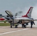 A United States Air Force Thunderbirds F-16 Fighting Falcon is on the tarmac with its canopy open, surrounded by ground crew personnel. Smoke or exhaust is visible near the rear of the aircraft, suggesting engine startup or shutdown procedures are underway. At least two additional Thunderbirds aircraft are partially visible on either side of the primary aircraft.